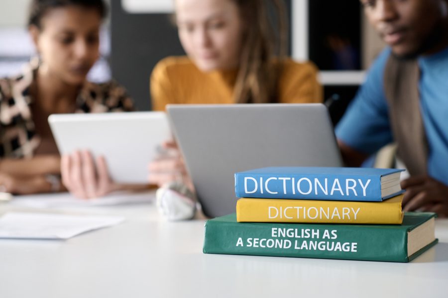 Close up background image of stacks of books on table with students studying in background, copy space