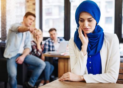 Multicultural society. Pleasant beautiful young muslim woman sitting at the table in the cafe while her mates abusing her