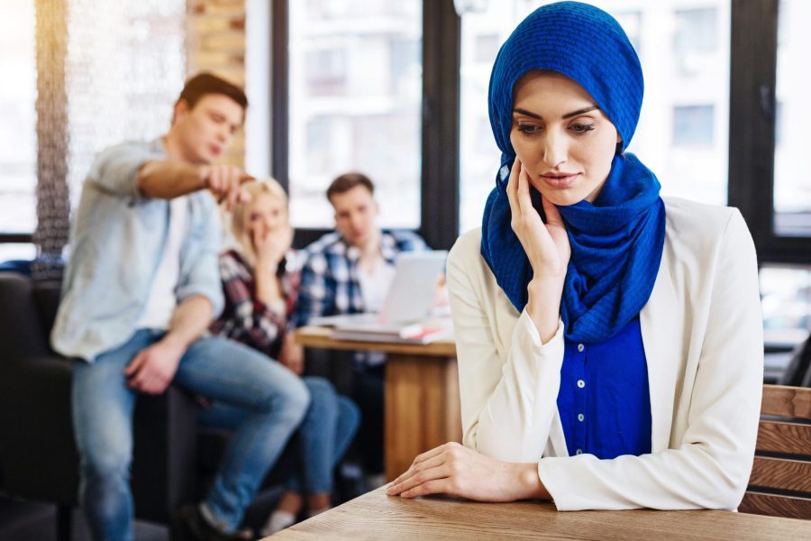 Multicultural society. Pleasant beautiful young muslim woman sitting at the table in the cafe while her mates abusing her